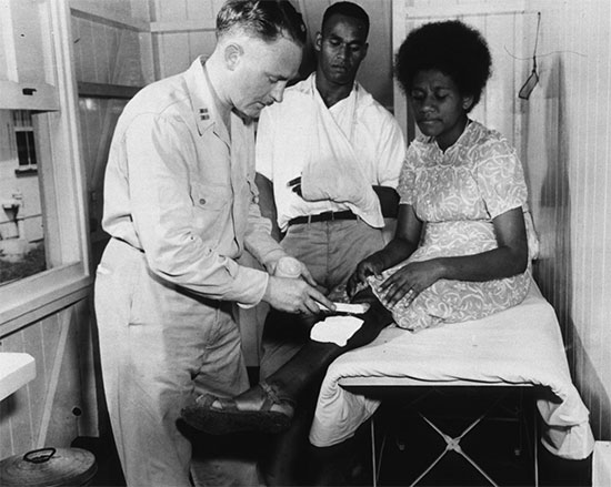  At the Out-Patient Clinic of the 71st Station Hospital in Fiji, Capt. George Warnock, MC, of Freeport, LI, treats a Fijian woman, Stery Uquaqe, for burns, as her husband, Roney, looks on.