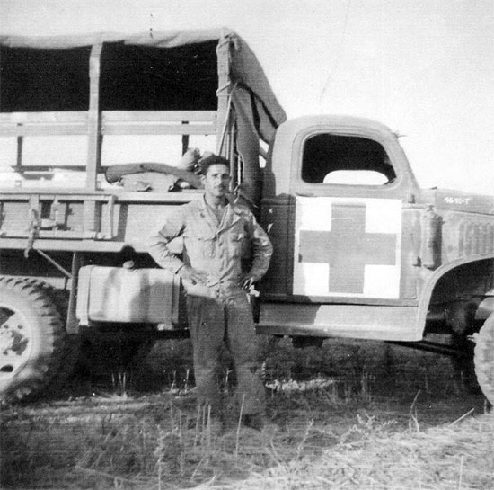 Driver J. D. Lloyd poses for the camera in front of a 2 1/2-ton Truck assigned to the 11th Evacuation Hospital. Of interest is the large Geneva Convention markings which have been applied to the door of the vehicle. 